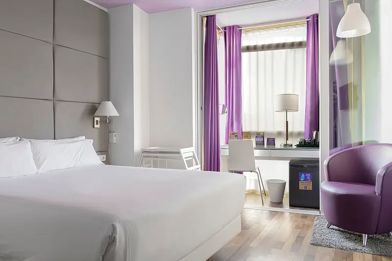 Habitación doble con cama grande, sillón morado y escritorio junto al ventanal
