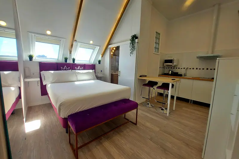 Estudio abuhardillado con cama, cabecero morado con coronas y cocina americana