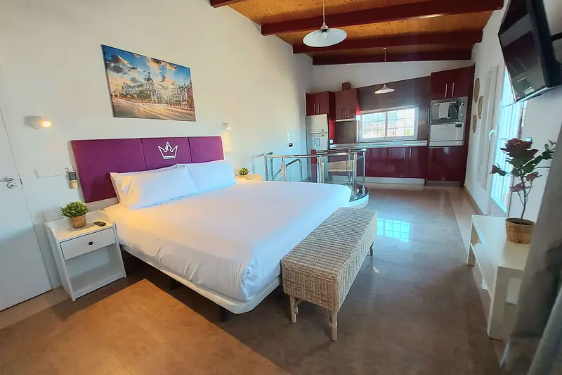 Estudio con techo de vigas de madera, cama con cabecero morado y cocina roja al fondo