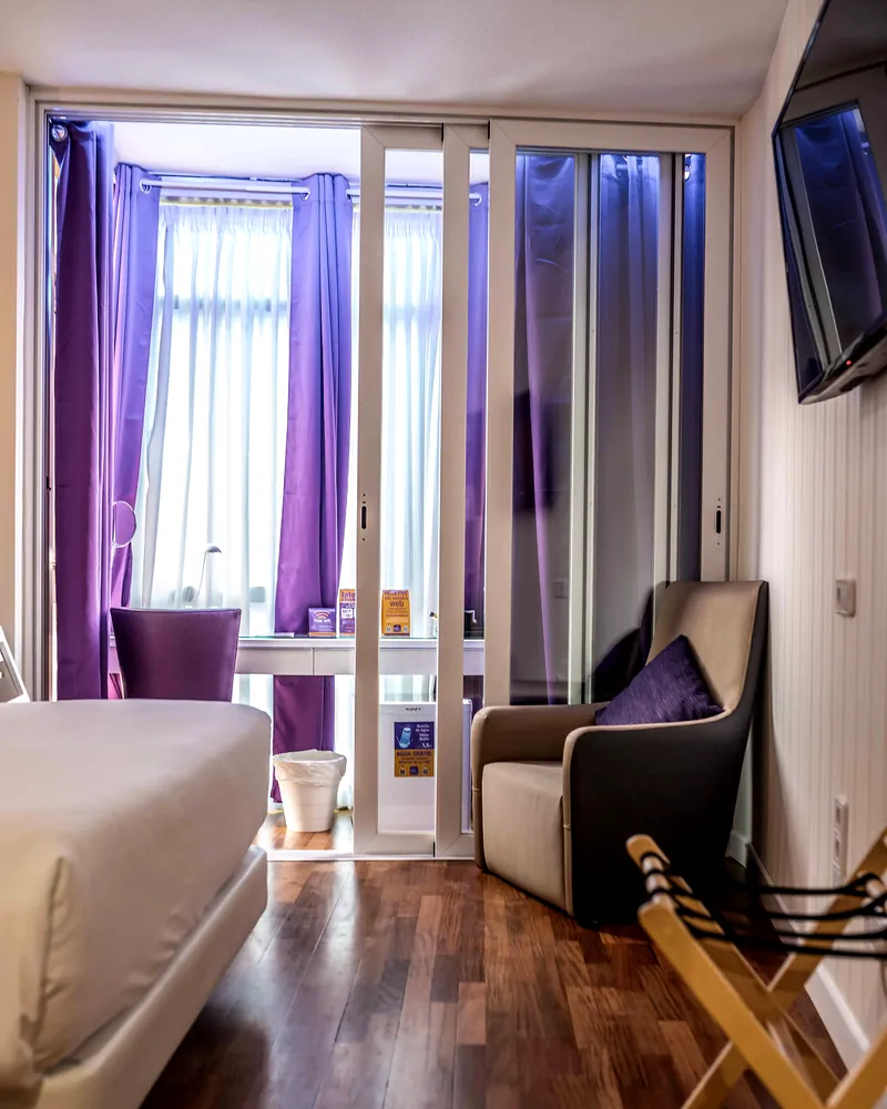 Habitación con cama, butaca y ventanal con cortinas moradas